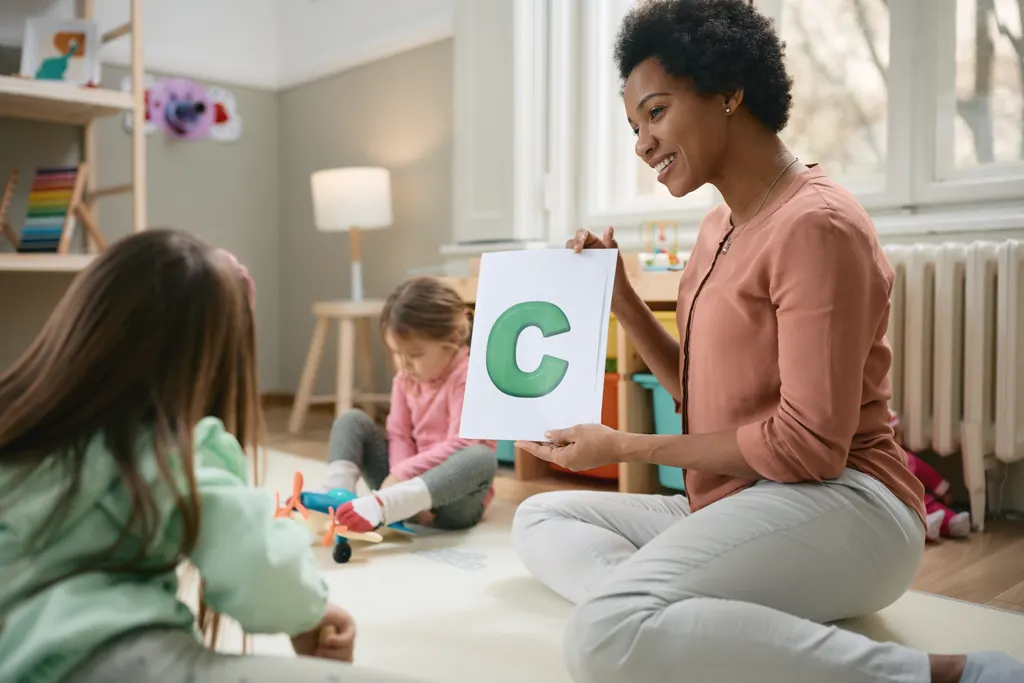 A smiling practitioner sits on the play mat by the window and holds up a big green letter C card, while a little girl plays with a toy aeroplane behind her