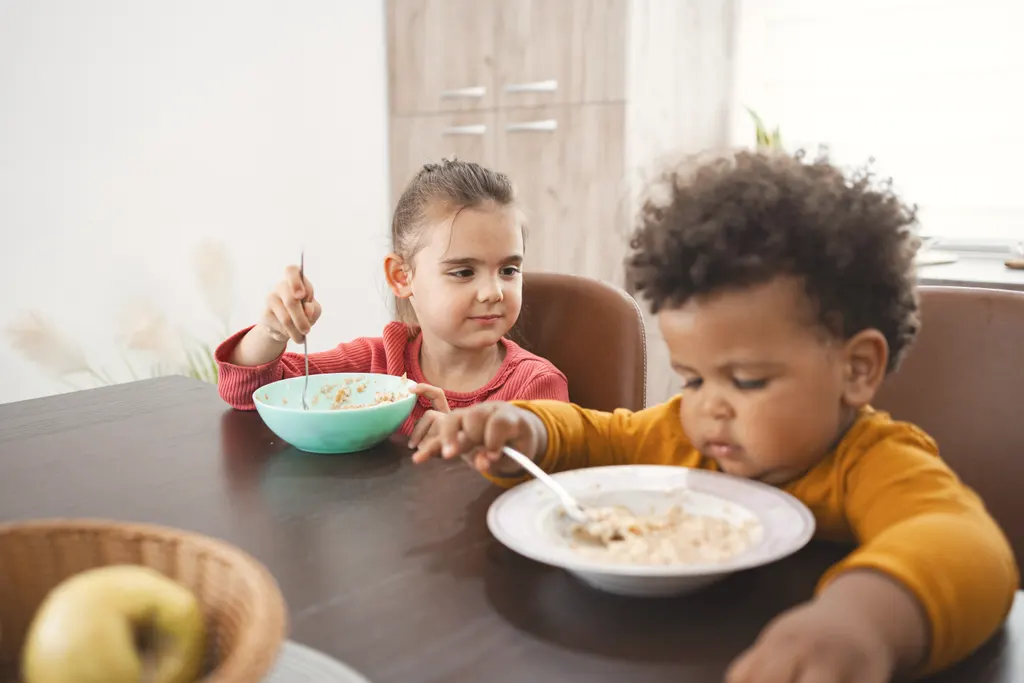 Two toddlers eating porridge at a wooden table with an apple nearby